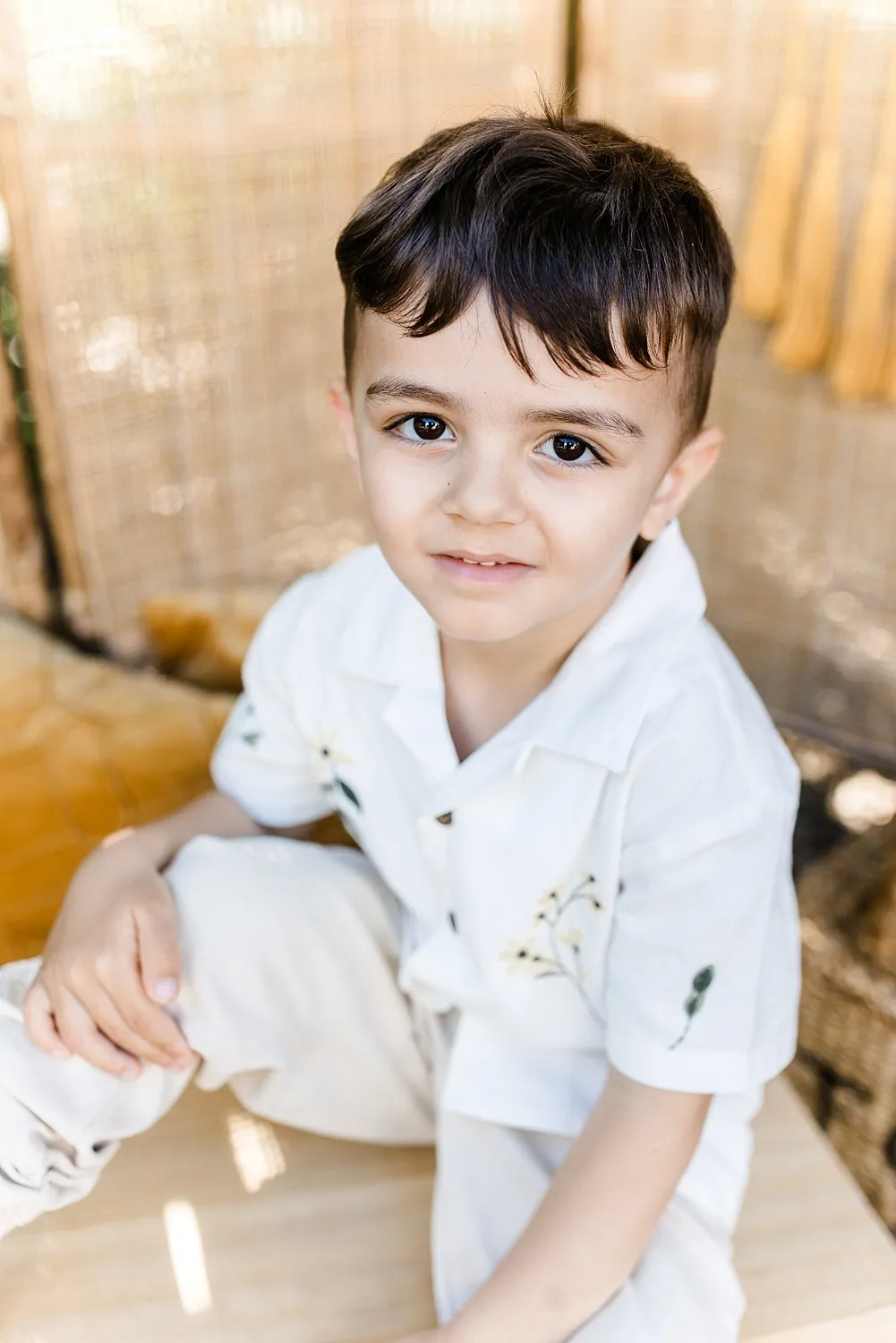 Kleiner Junge mit dunklen Haaren trägt helle Kleidung und sitzt vor einem geflochtenen Hintergrund.