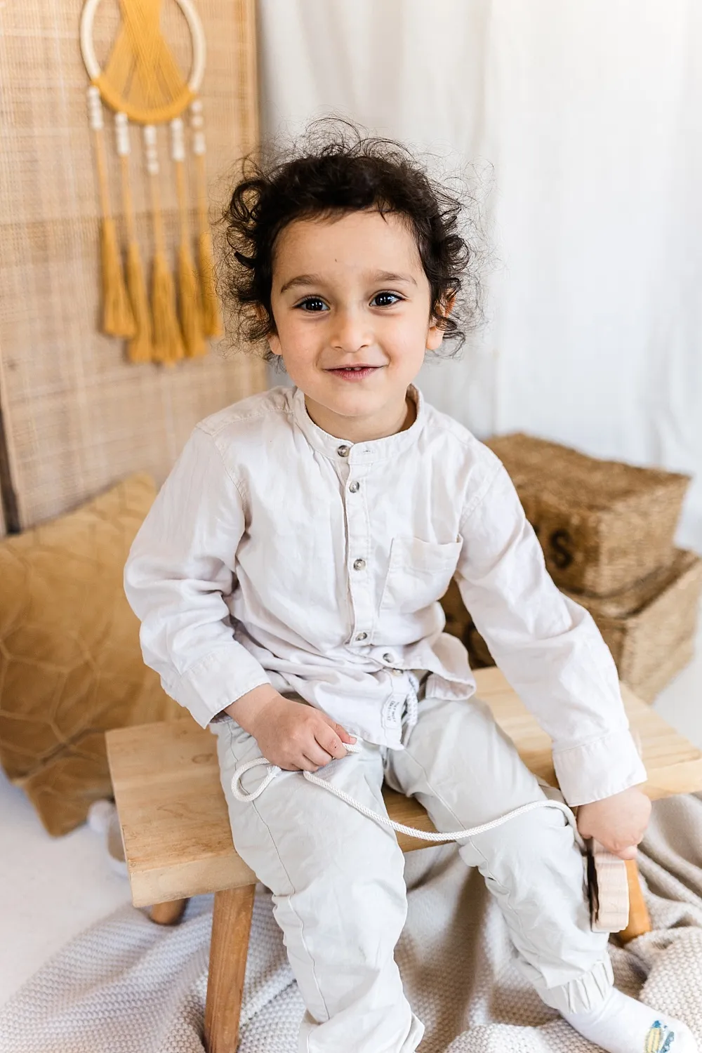 Kleines Kind mit lockigem Haar sitzt auf einer Holzbank und hält ein Spielzeug mit Schnur.