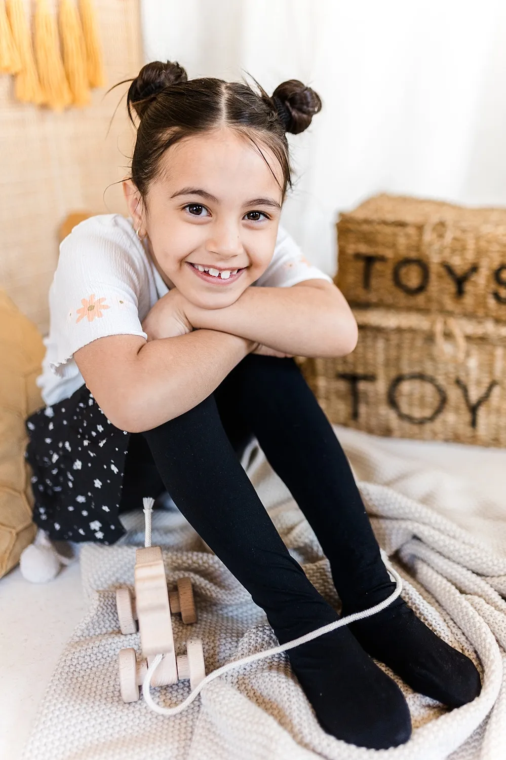 Lächendes Mädchen mit Zöpfen sitzt auf einer Decke neben einem Holzspielzeugauto und zwei Körben mit der Aufschrift 'TOYS'.