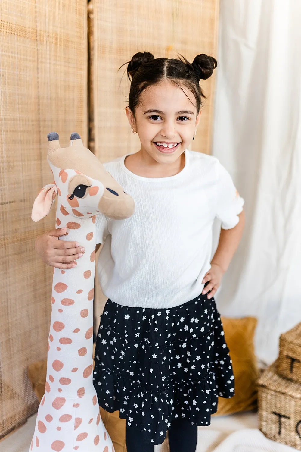 Mädchen mit weißen Shirt und schwarzem Rock hält ein großes Plüschgiraffen-Spielzeug und lächelt.