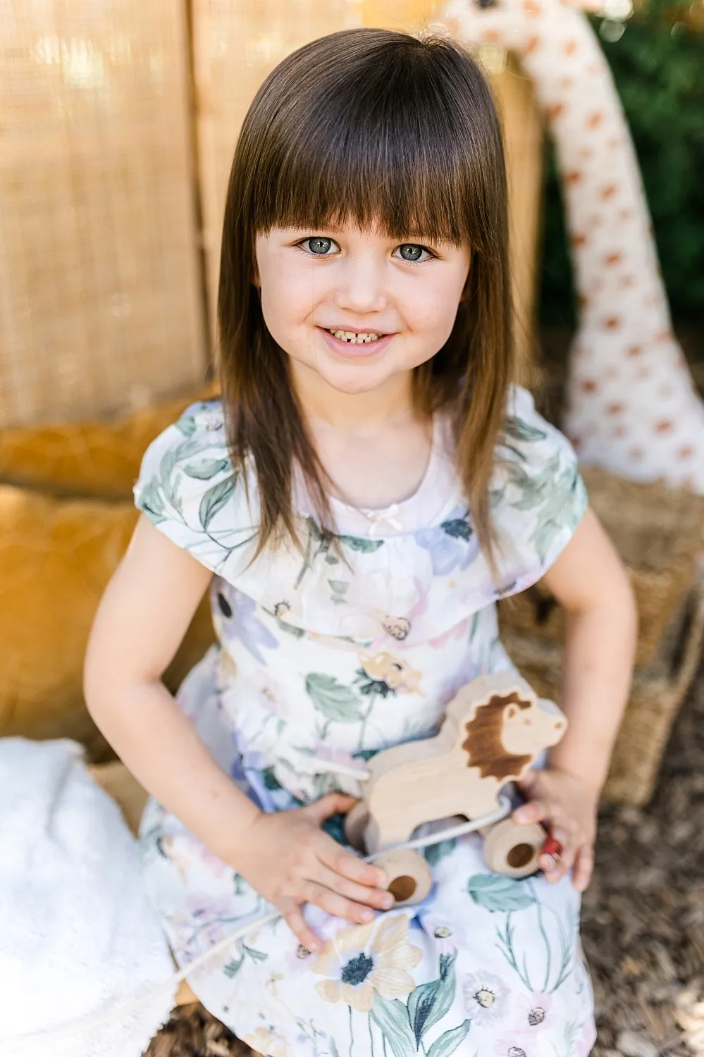 Lächendes kleines Mädchen mit braunen Haaren und floralem Kleid hält ein Holzspielzeug in Form eines Löwen.