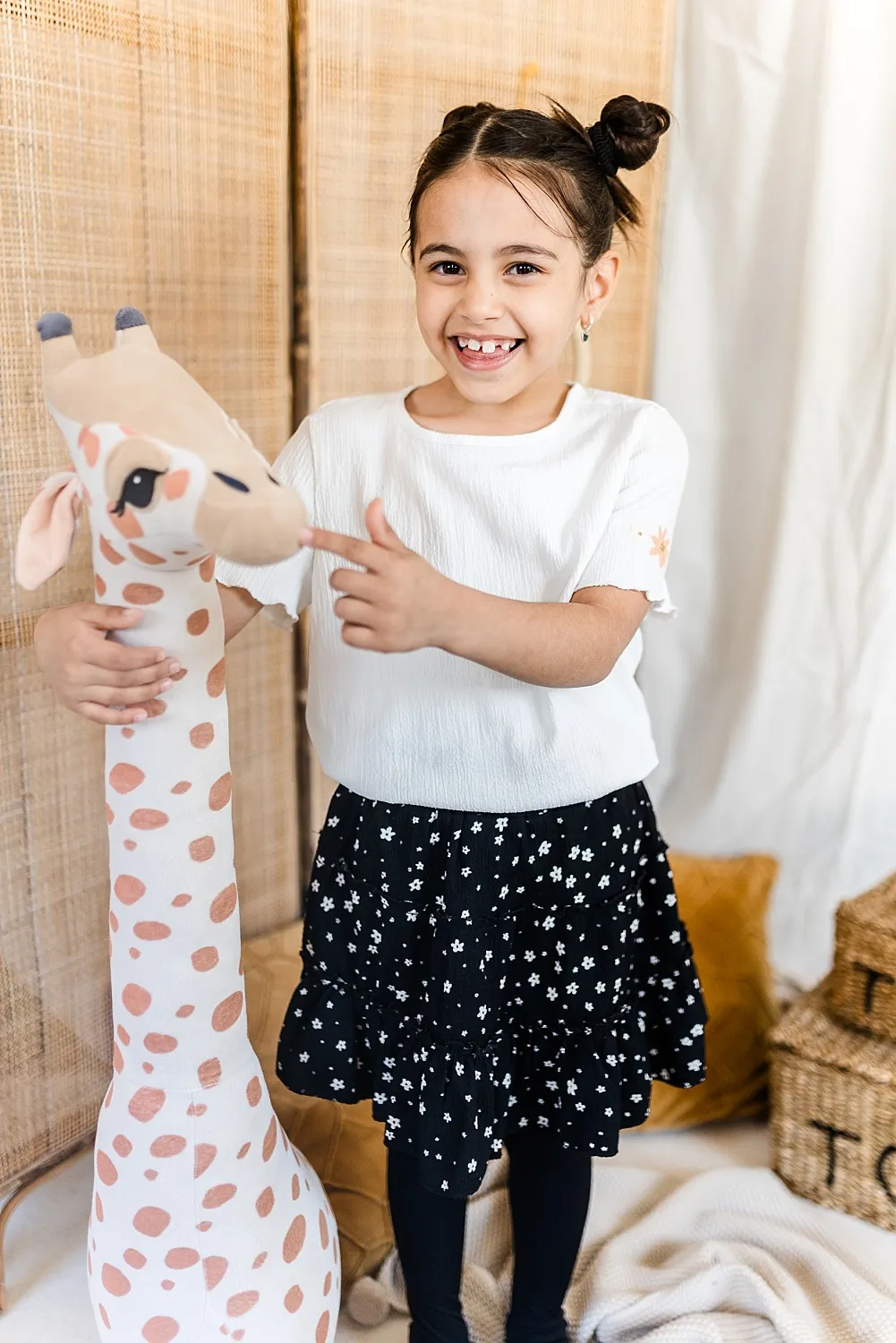 Fröhliches Mädchen mit Zahnlücke in weißem Oberteil und schwarzem Rock zeigt auf ein großes Plüschgiraffen-Spielzeug.