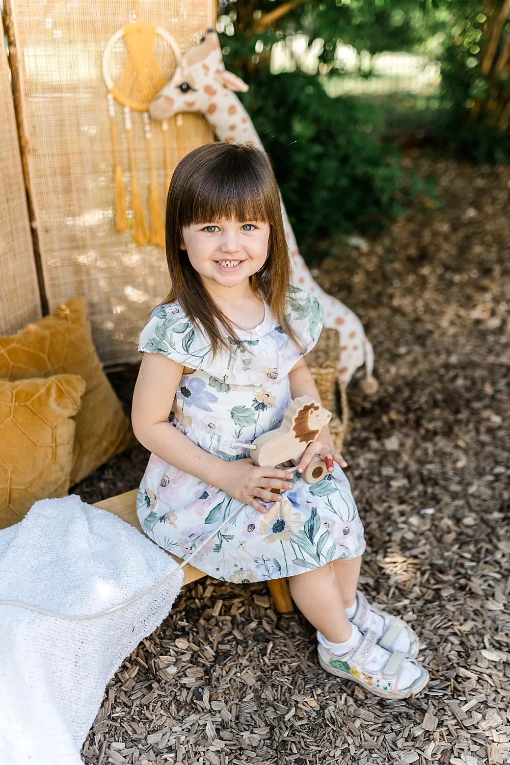 Kleines Mädchen mit Blumenkleid sitzt draußen auf einem Holzstuhl und hält ein hölzernes Löwenspielzeug.