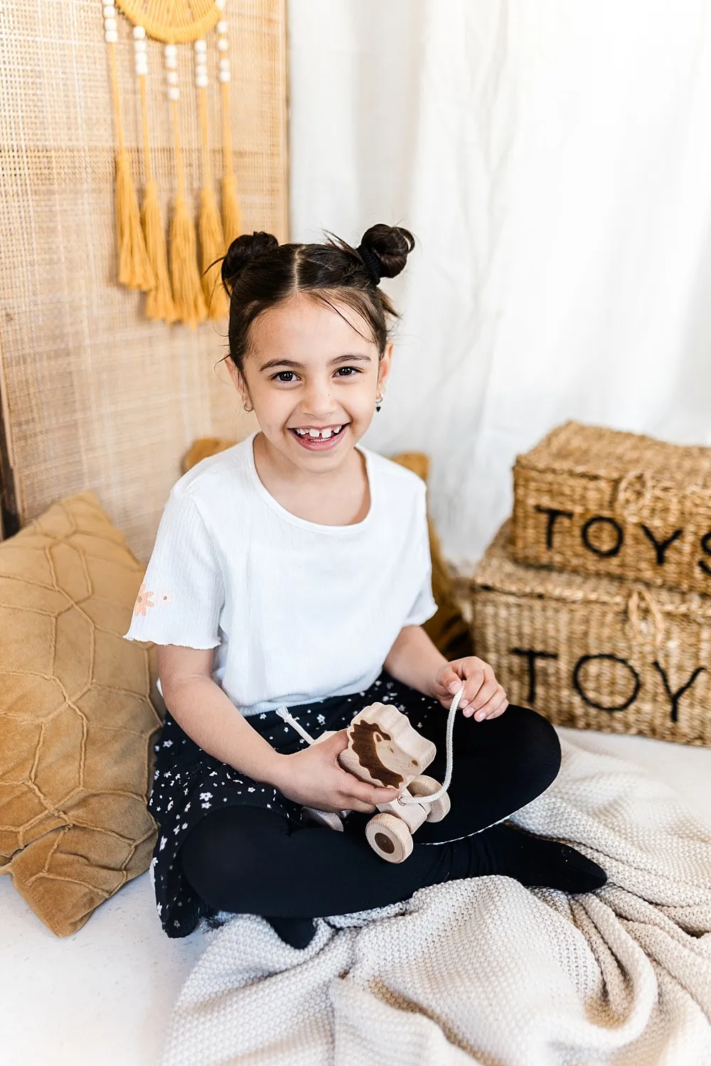 Lächendes Mädchen mit zwei Duttfrisuren sitzt auf einer Decke und hält ein hölzernes Spielzeuglöwenfahrzeug.