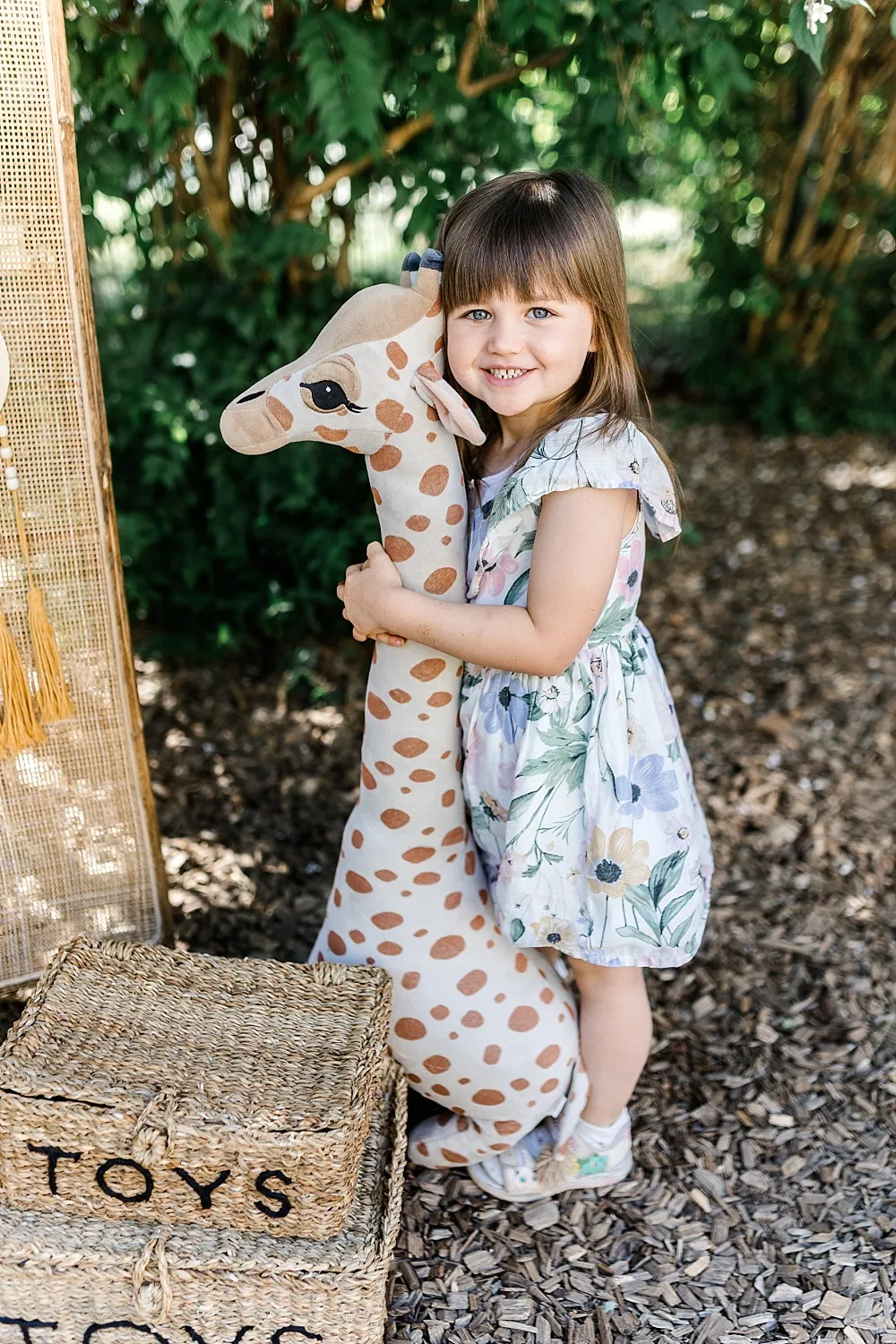 Kleines Mädchen in geblümtem Kleid umarmt ein großes Stoffgiraffen-Spielzeug im Freien.