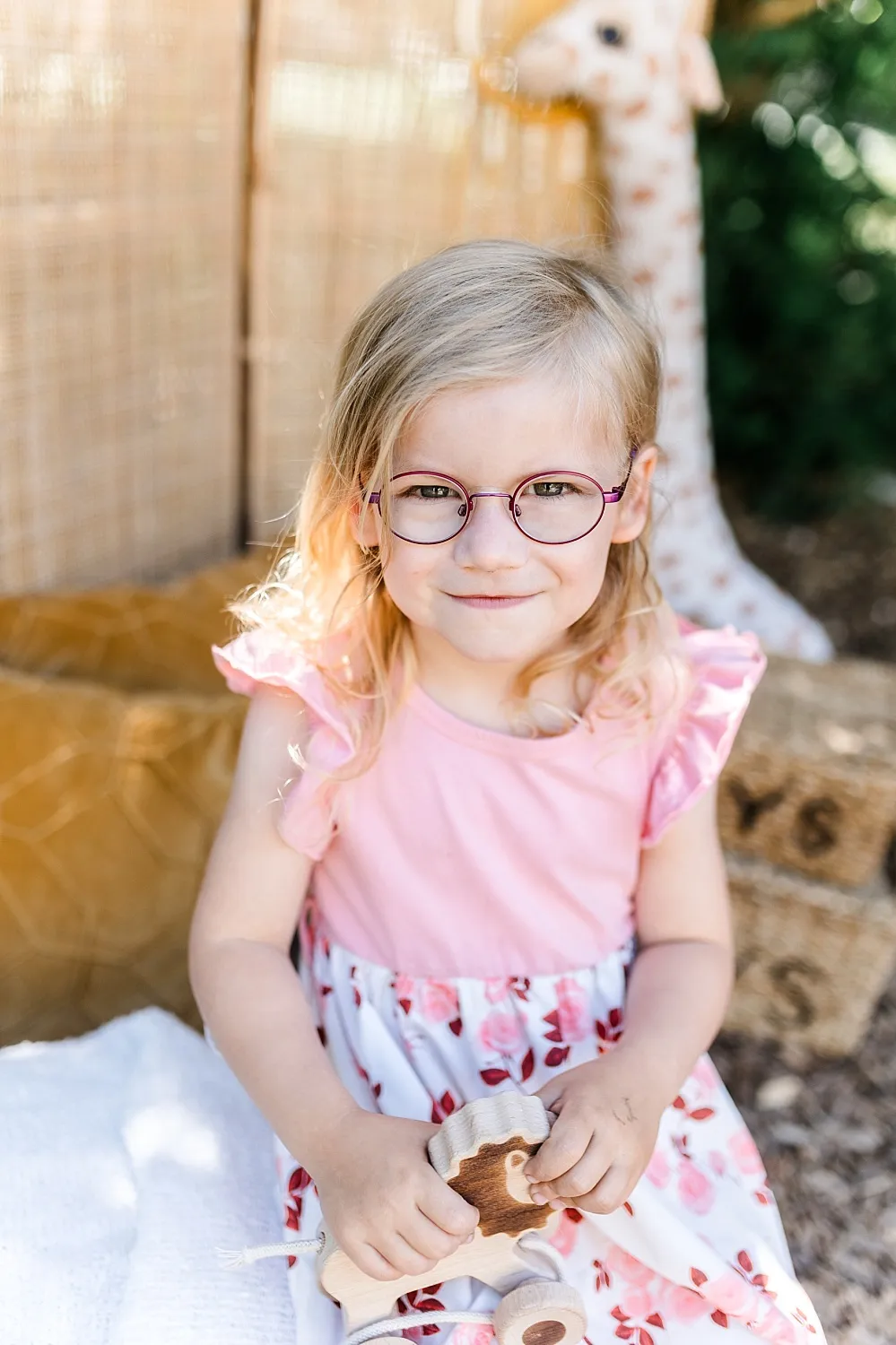 Kleines Mädchen mit Brille in rosa Kleid hält ein Holzspielzeug, sitzend vor einer geflochtenen Wand und einem Stoffgiraffen-Spielzeug.