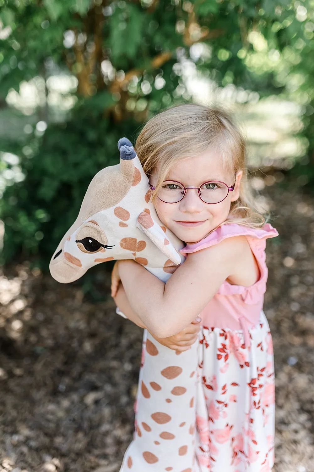 Kleines Mädchen mit Brille umarmt eine große Plüschgiraffe im Freien.