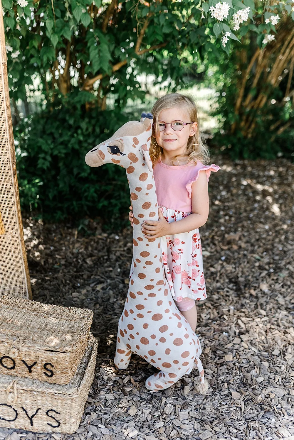 Kleines Mädchen mit Brille huggt ein großes, geflecktes Stoffgiraffen-Spielzeug im Garten.