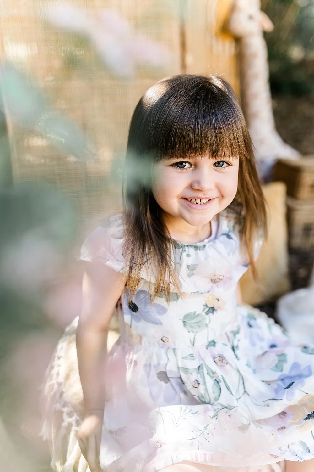 Kleines Mädchen mit braunen Haaren und Blumenmuster-Kleid lächelt im Sonnenlicht.