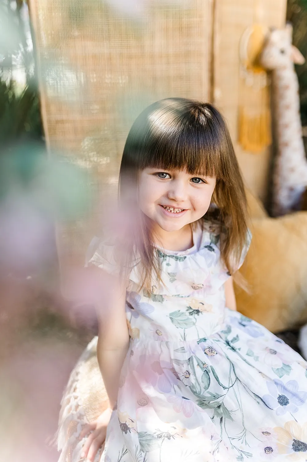 Kleines Mädchen mit braunen Haaren und Pony in einem weißen Kleid mit Blumenmuster lächelt in die Kamera.