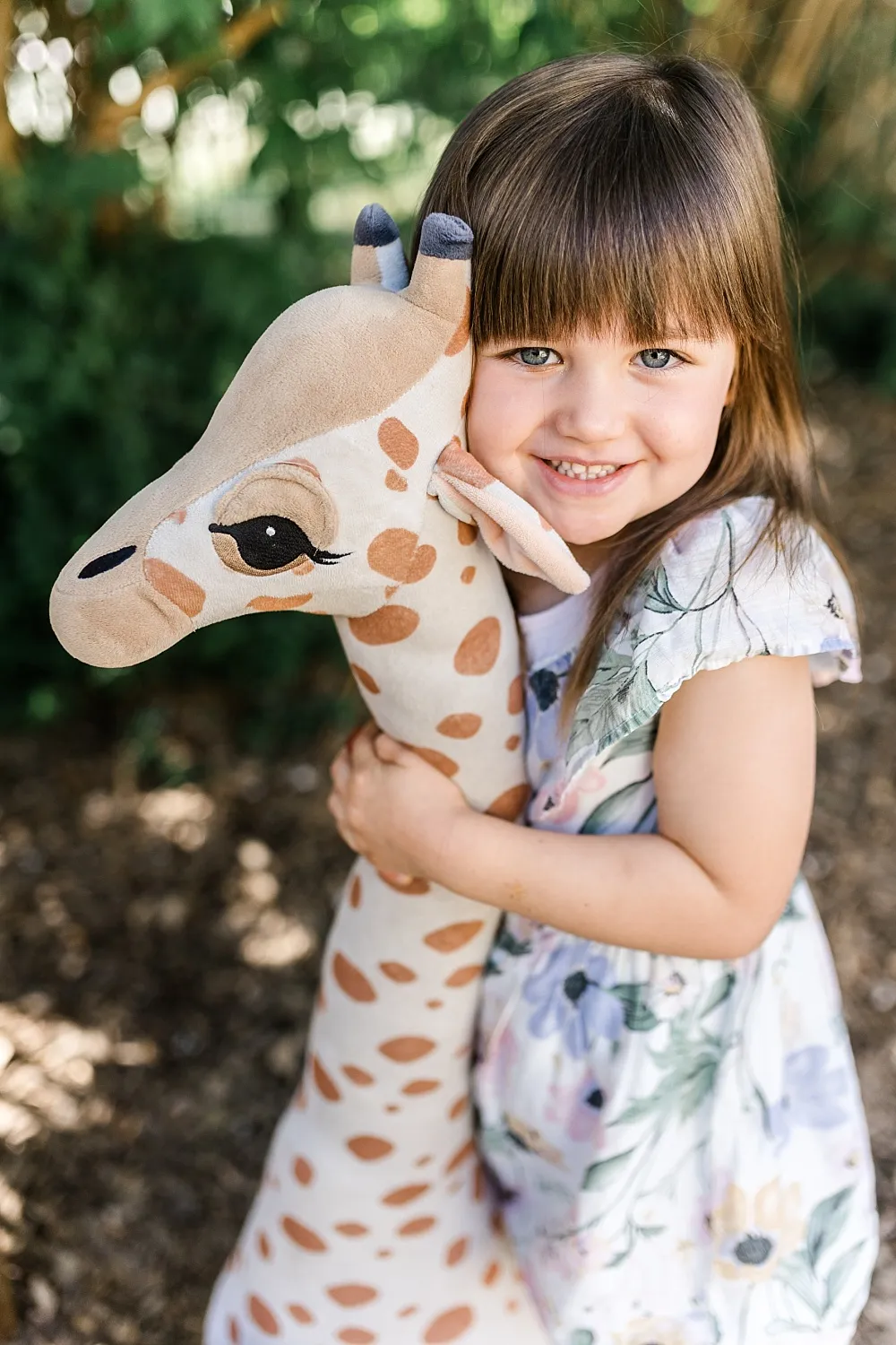 Kleines Mädchen mit Kleid umarmt lachend ein großes Stoffgiraffen-Spielzeug im Freien.