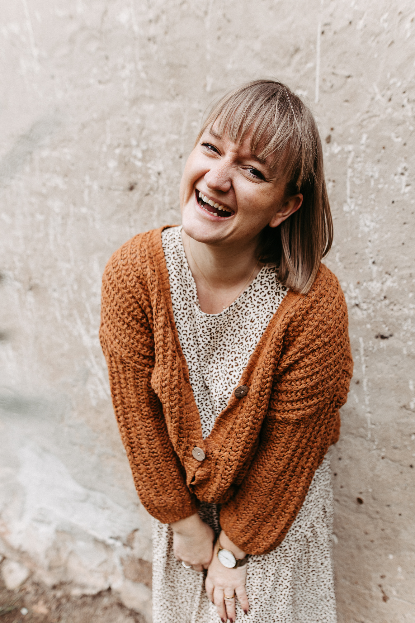 Lächelnde Frau mit kurzem blonden Haar, brauner Strickjacke und weißem Kleid mit Muster, die draußen auf einem Baumstamm sitzt und eine Kamera hält.