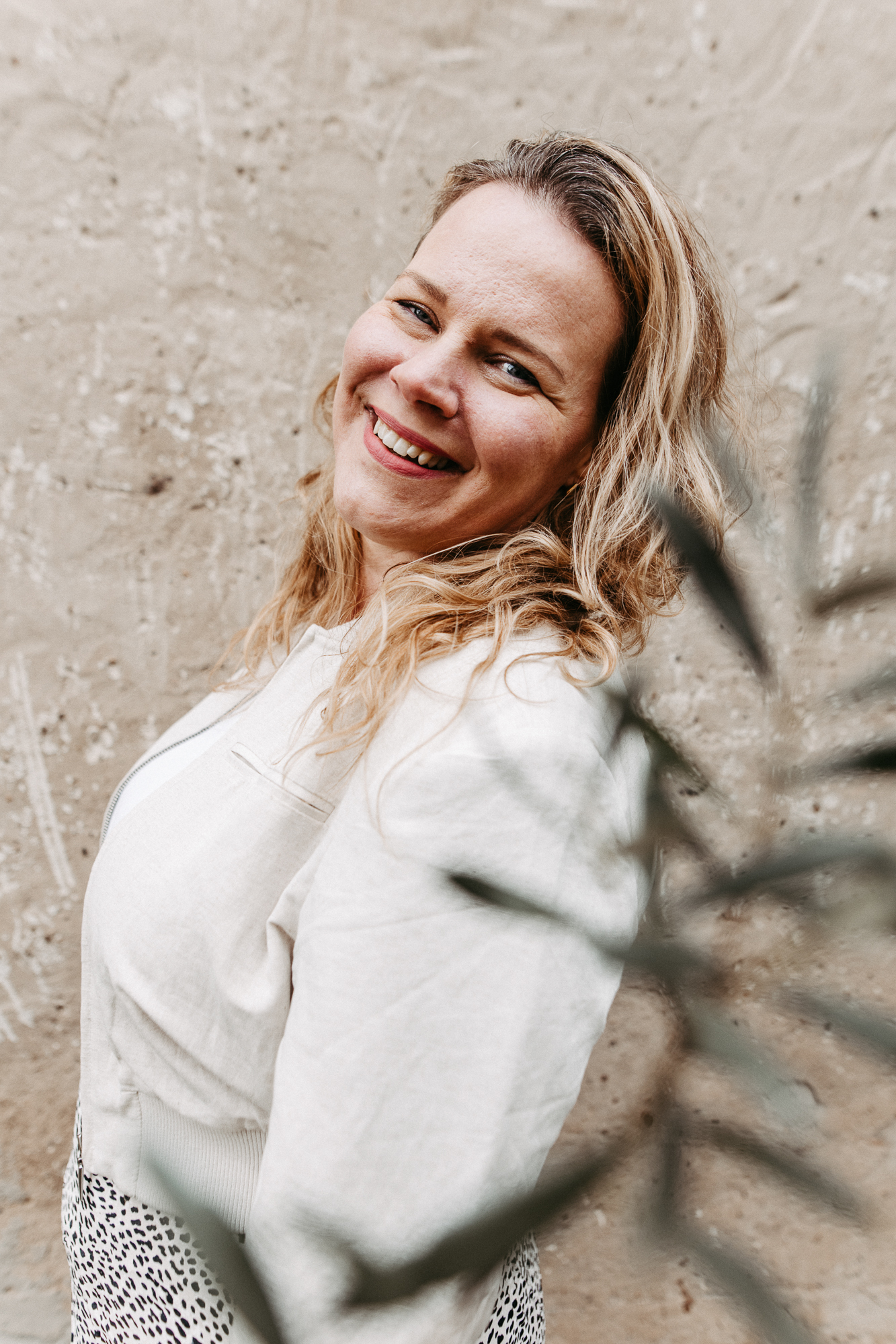 Lächelnde Frau mit langem, welligem Haar, weißer Bluse und blauer Jeans steht in einem sonnigen Wald.