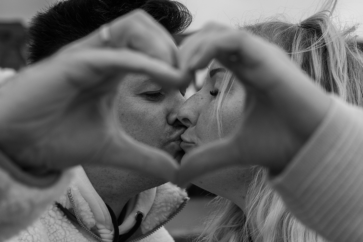 Ein Paar küsst sich. Im Vorergrund formen sie ihre Hände zu einem Herz.