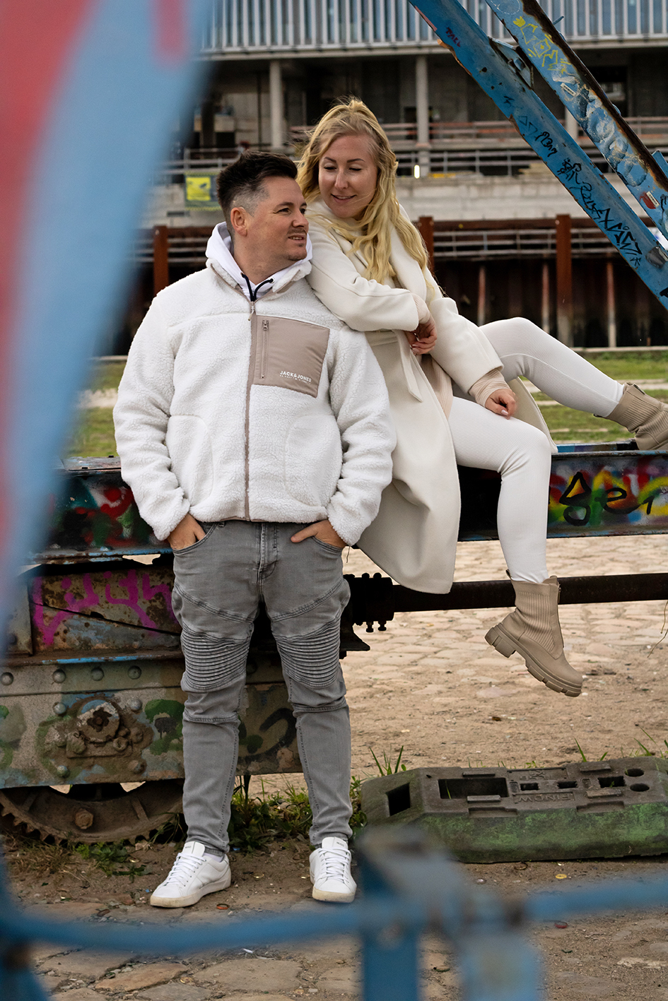 Zu sehen ist ein Paar. Sie sitzt auf einem Geländer und schaut zu ihm. Er steht am Geländer angelehnt und schaut in die Ferne.