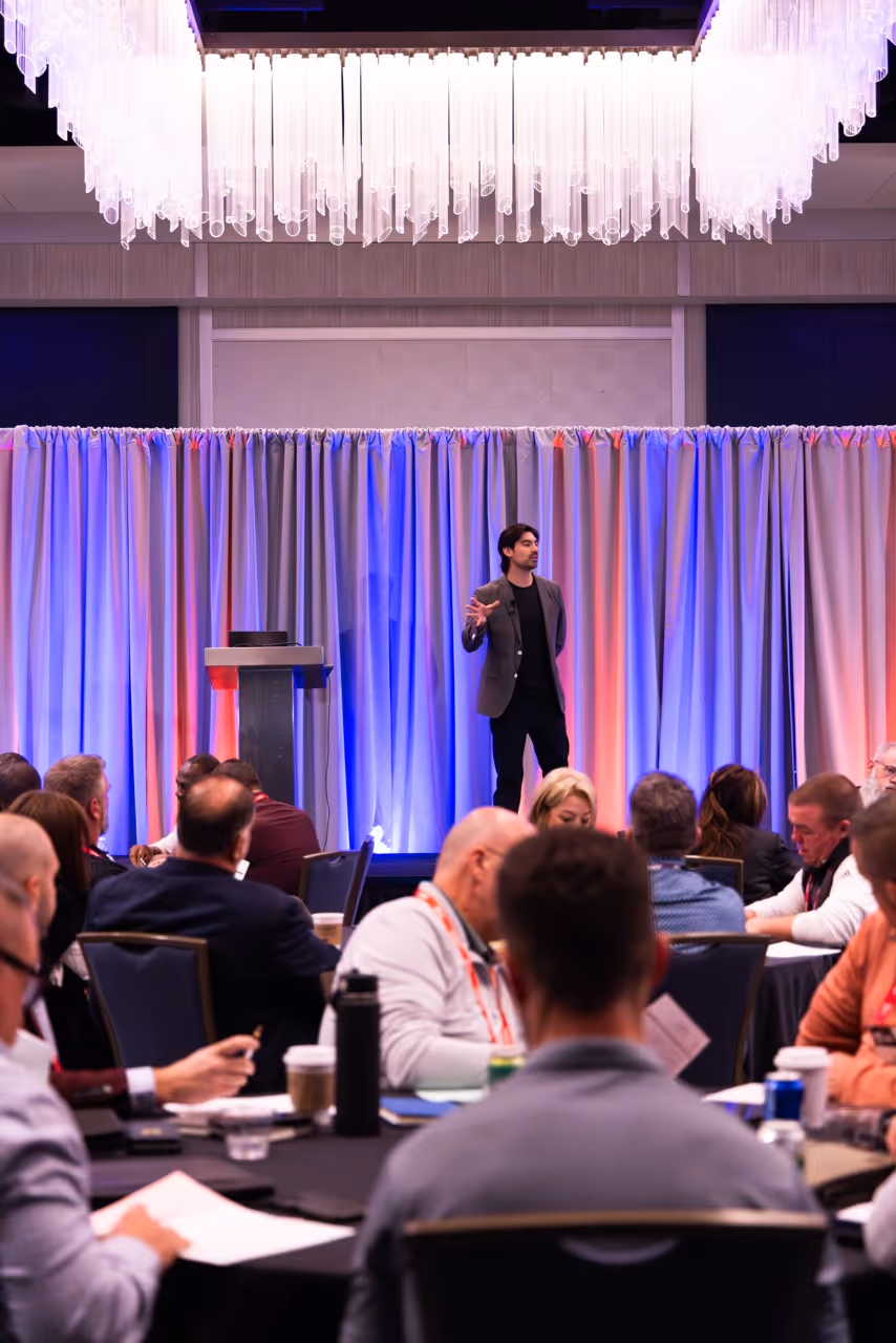 Speaker presenting on stage with gray curtains and audience seated at round tables in a conference room.