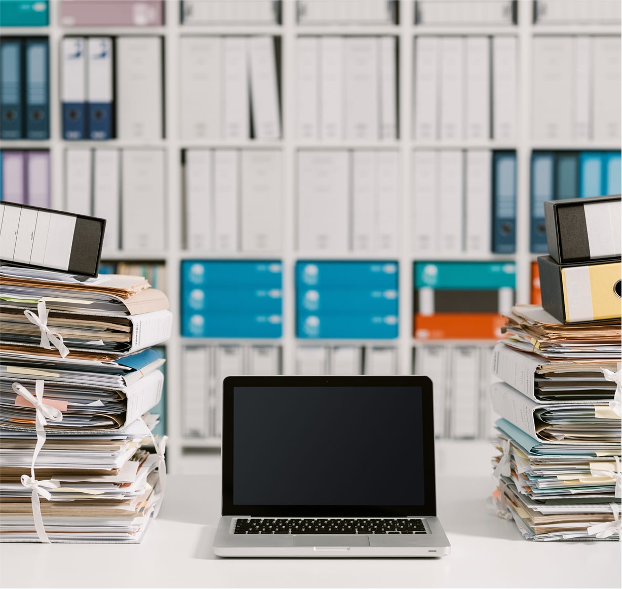 Laptop on desk surrounded by large stacks of paper files