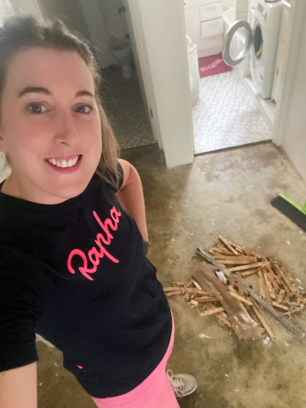 Woman smiling in a black shirt and pink pants standing on a bare floor with a pile of wooden debris near a washing machine in a laundry area.