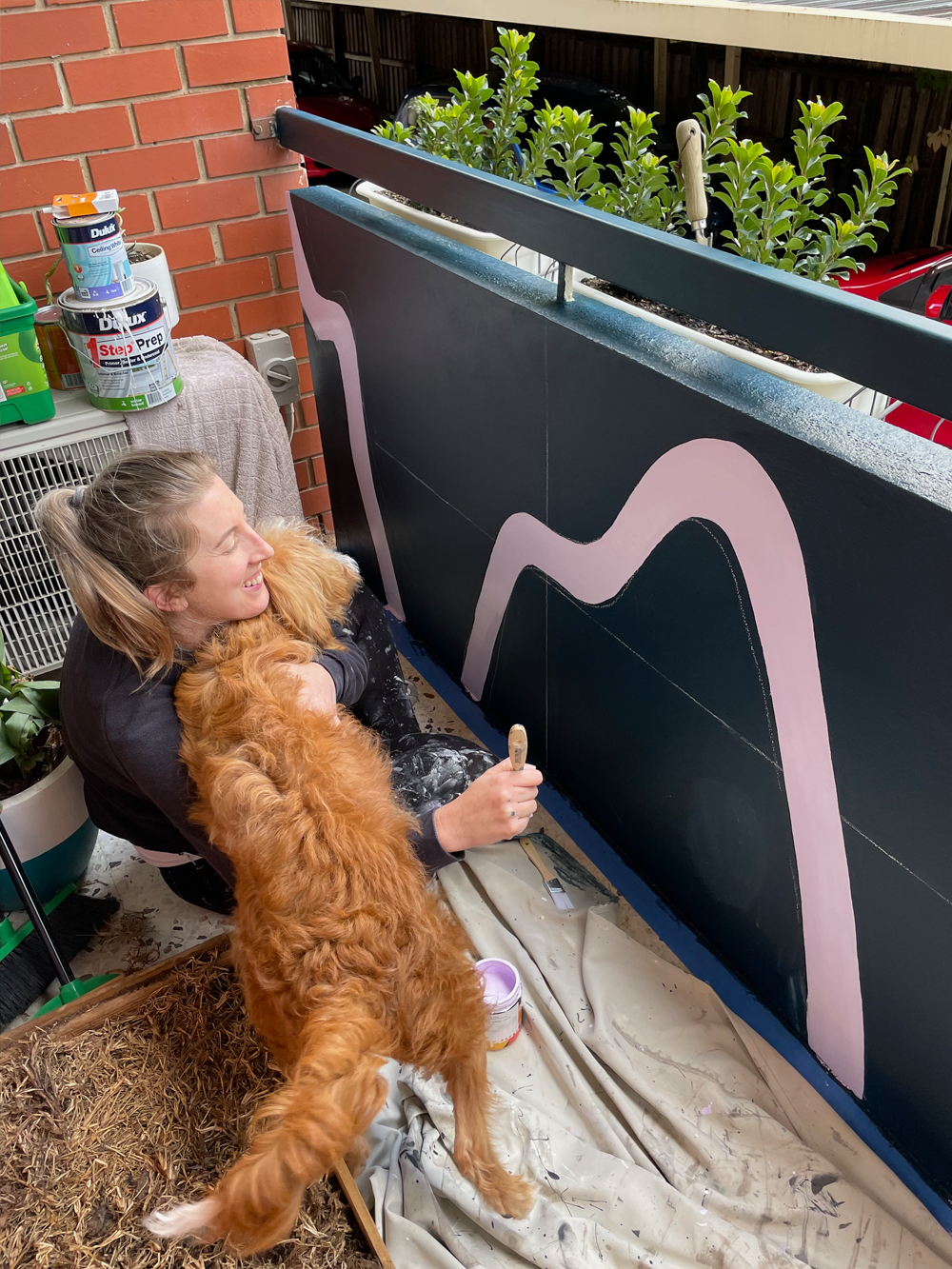 Woman sitting on the floor hugging a large fluffy brown dog next to a painted balcony railing with pink abstract shapes.