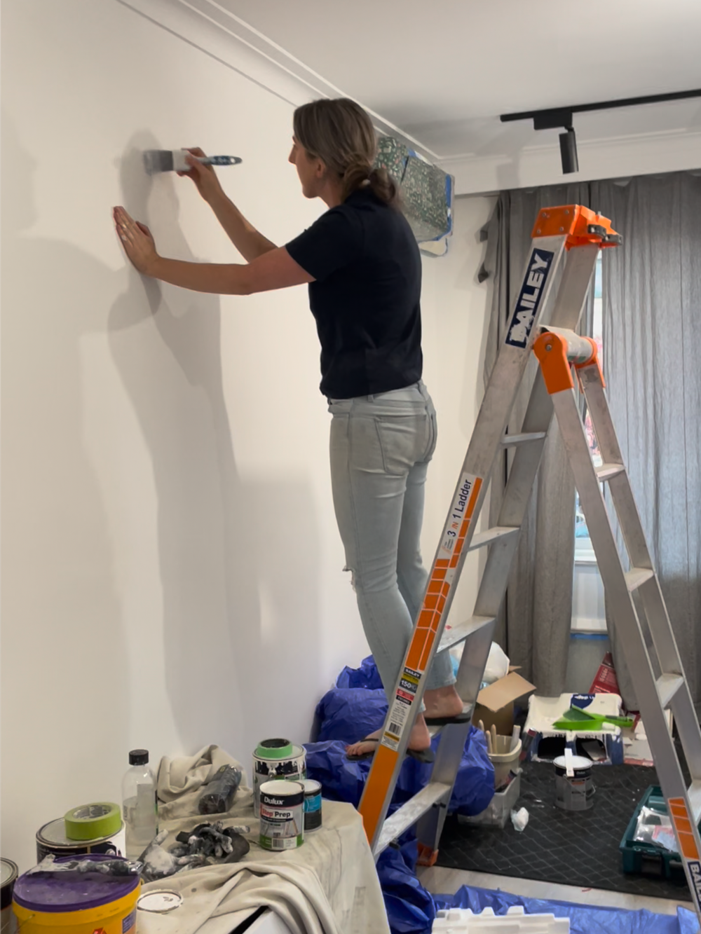 Woman standing barefoot on a ladder painting a white wall with a paintbrush in a room with painting supplies on a covered table.