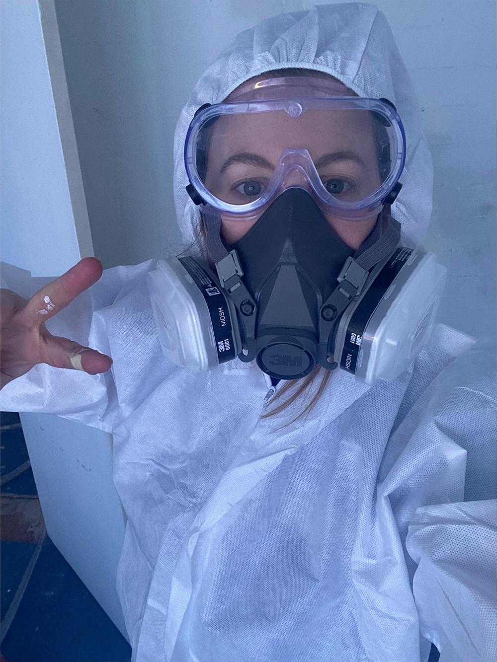 Woman wearing a white protective coverall, clear safety goggles, and a black 3M respirator mask showing a peace sign with their hand.