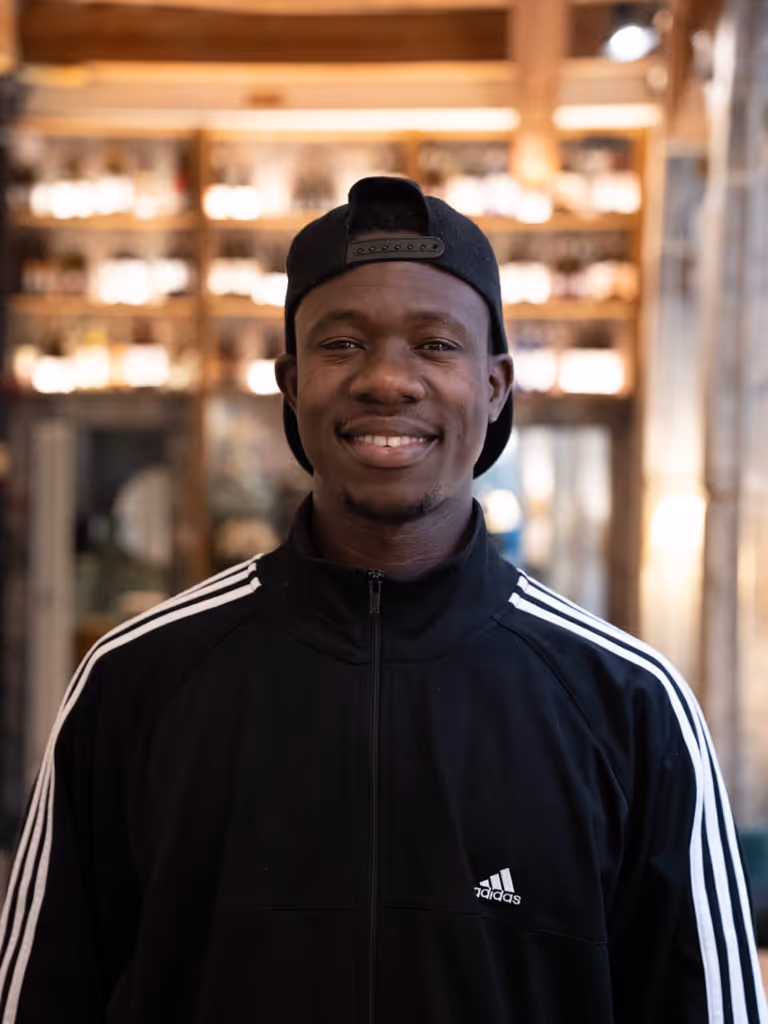Hombre joven sonriente con gorra negra al revés y chaqueta deportiva Adidas negra con rayas blancas en interiores de edificio.