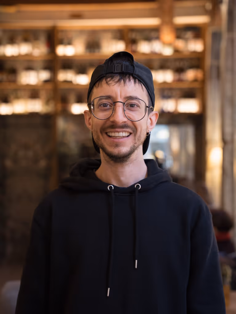 Hombre joven con gafas redondas, gorra negra al revés y sudadera negra sonriendo en interior con estanterías iluminadas al fondo.