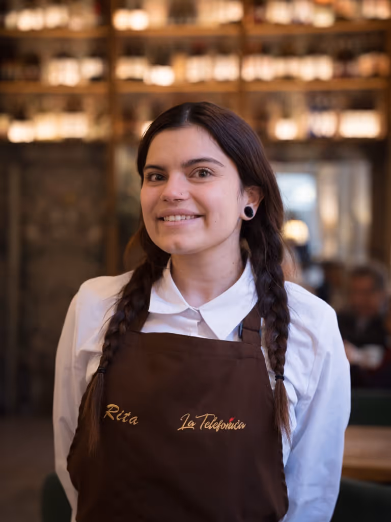 Mujer joven con trenzas y delantal marrón que dice 'Rita La Telefónica' sonriente en un ambiente de restaurante.