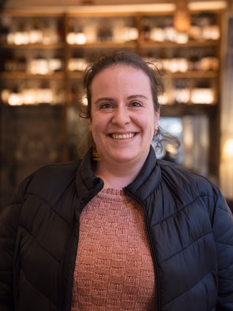 Mujer sonriente con chaqueta negra y suéter rosa en un ambiente interior con estantes iluminados al fondo.