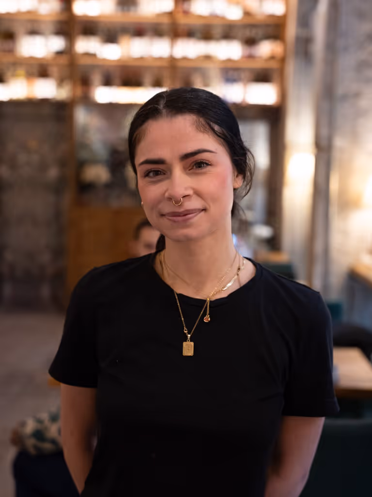 Mujer joven con cabello oscuro recogido, vestida con camiseta negra y dos collares dorados, sonriendo ligeramente en un entorno interior desenfocado.