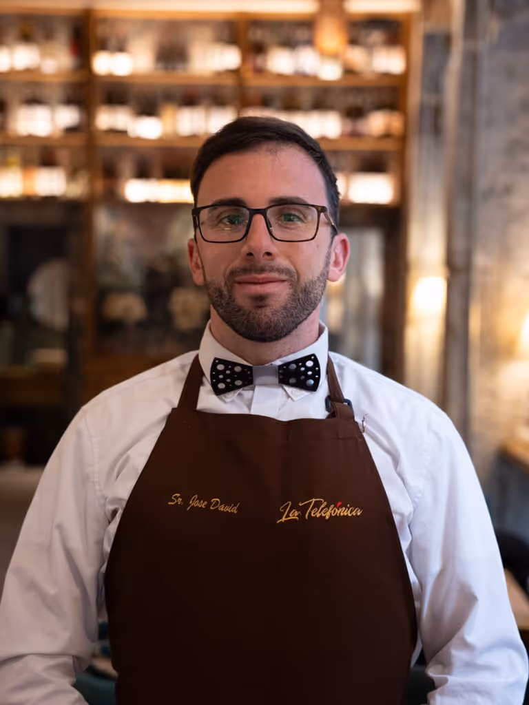Hombre con gafas, pajarita negra con puntos blancos y delantal marrón con el nombre 'Sr. Jose David' y 'La Telefónica' en un restaurante.