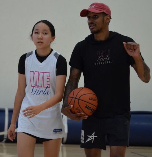 A young female basketball player in a white WE Girls jersey stands next to a male coach holding a basketball, wearing a black WE Girls Basketball shirt and a red cap.