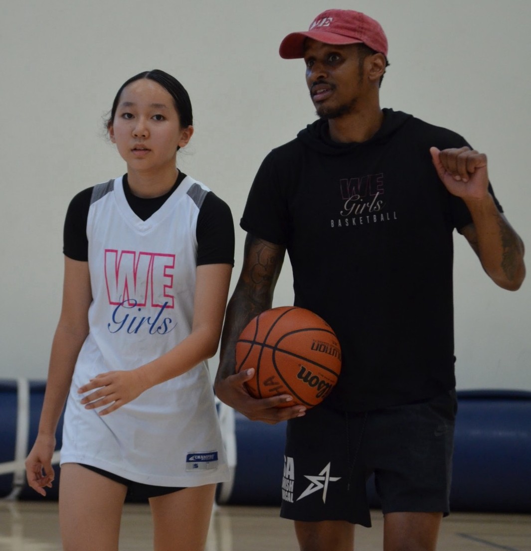 A young female basketball player in a white WE Girls jersey stands next to a male coach holding a basketball, wearing a black WE Girls Basketball shirt and a red cap.