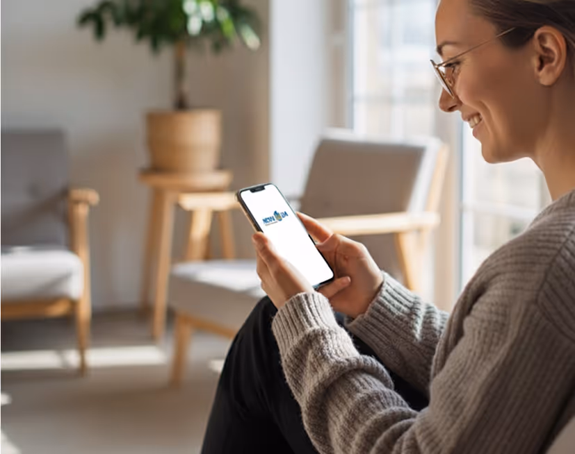 Smiling woman wearing glasses and a gray sweater looking at a smartphone in a bright living room.
