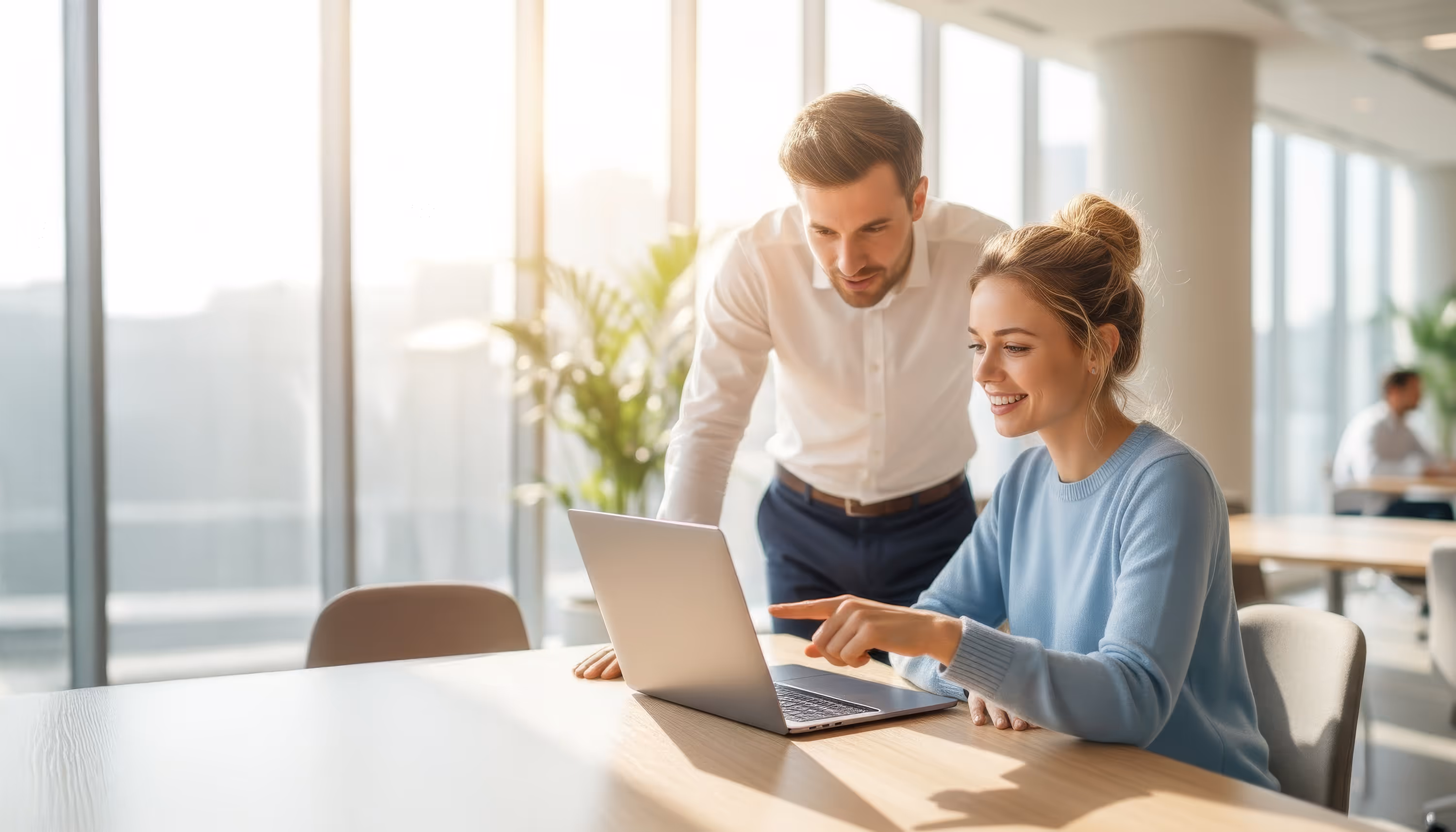 A smiling woman sitting at a desk and pointing at a laptop screen while a man in a white shirt stands beside her in a bright office.