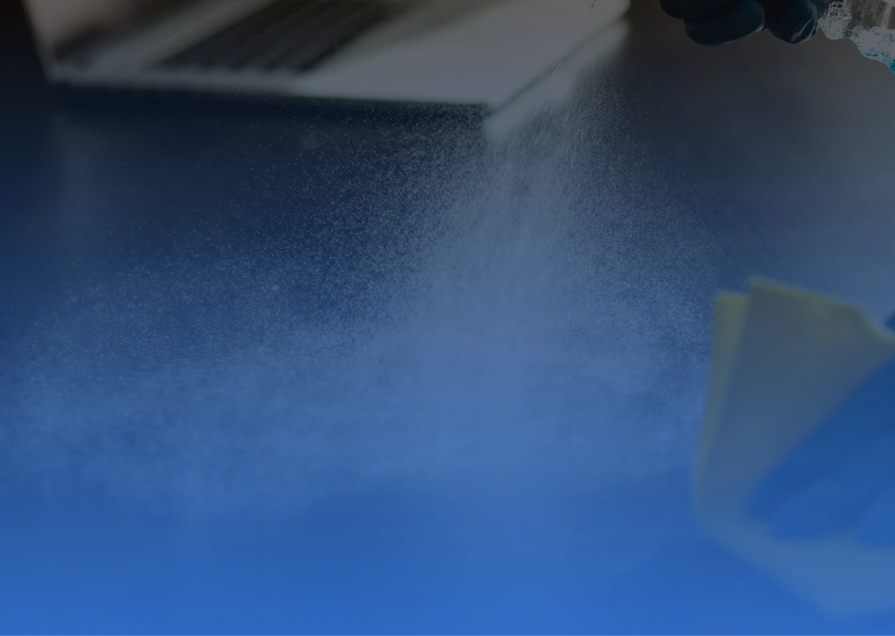 Hand mit Handschuh sprüht Reinigungsmittel auf eine Oberfläche mit Laptop im Hintergrund.