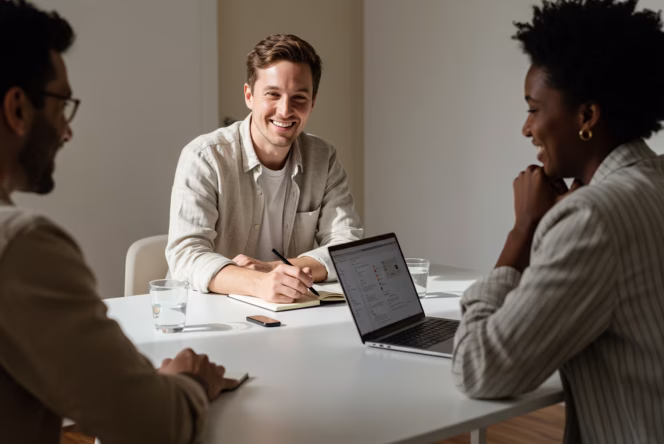 Three people having a friendly discussion around a white table with a laptop and notebooks.