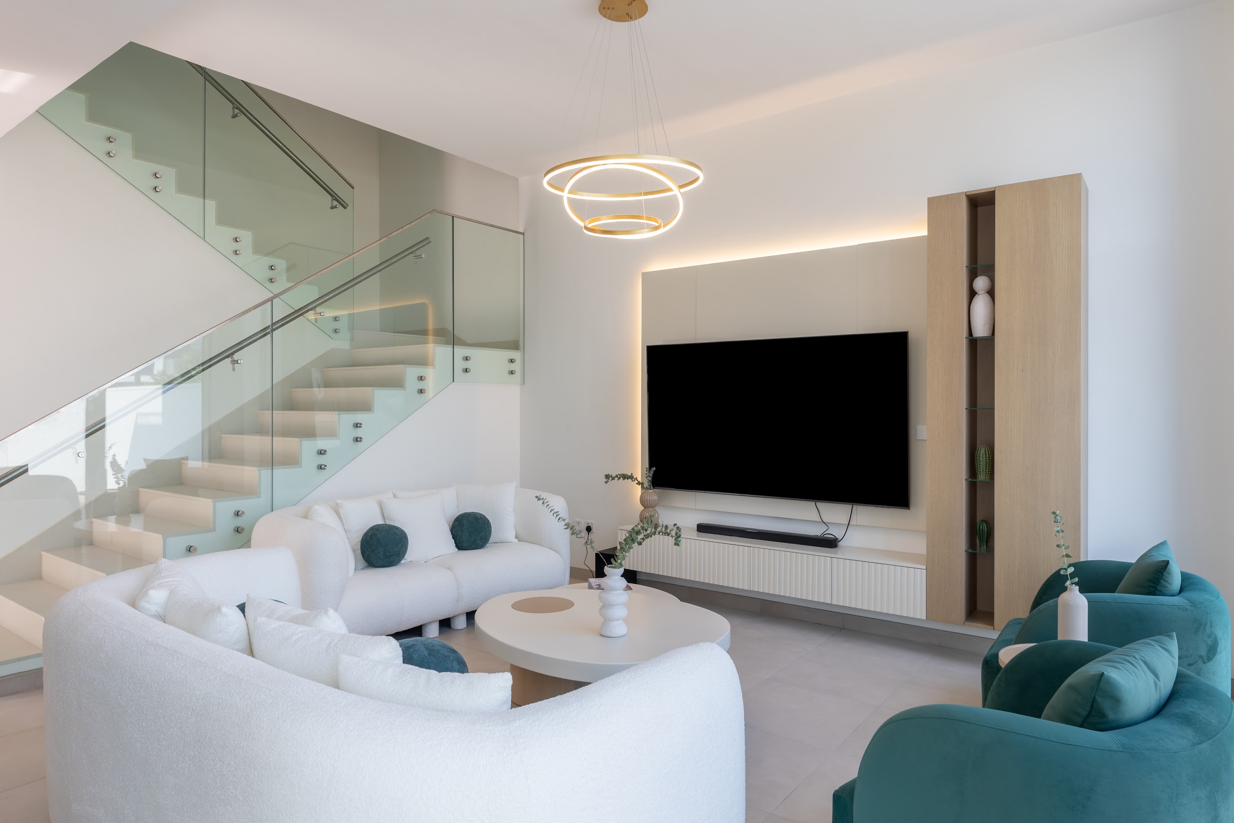 A cozy interior shot shows a black leather sofa next to a light wood open-shelf bench with a plant and books, below a light wood slatted staircase and a framed bird artwork.
