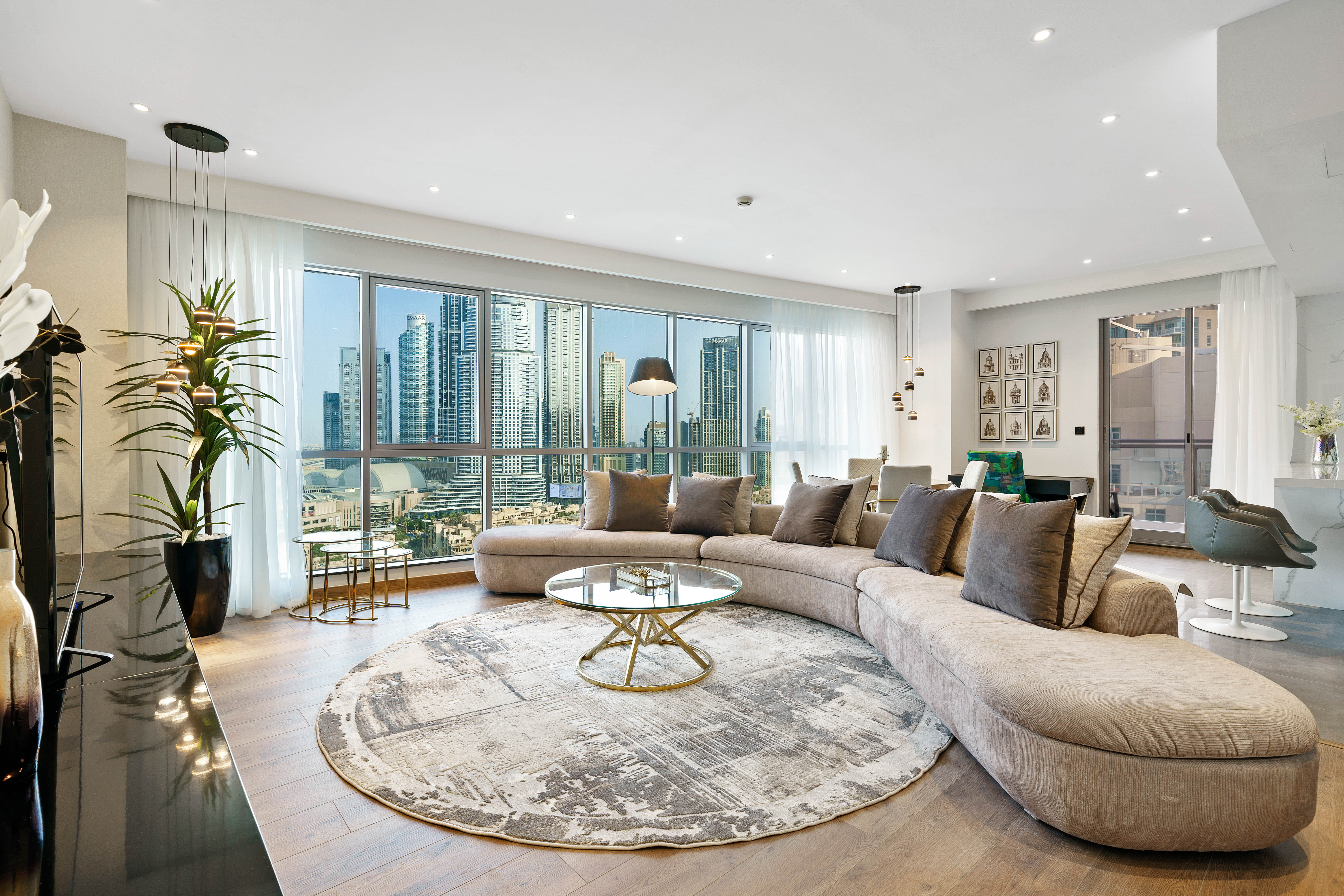 A spacious, minimalist room with light wood floors features a long dining table with black chairs in front of three tall windows, and a living area with a gray sofa and white coffee table on a patterned rug.