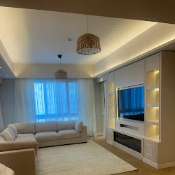 A luxurious bedroom showcases a white upholstered bed, gold chandelier, fuzzy white stools, and a black and white striped rug with a city view.