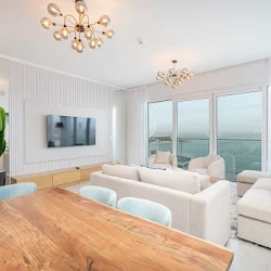 A luxurious bedroom showcases a white upholstered bed, gold chandelier, fuzzy white stools, and a black and white striped rug with a city view.