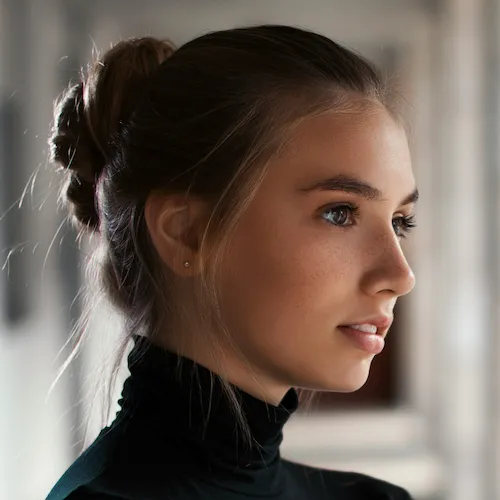 Profile of a real young woman with light brown hair in a bun, wearing a black turtleneck, softly lit with a blurred background.