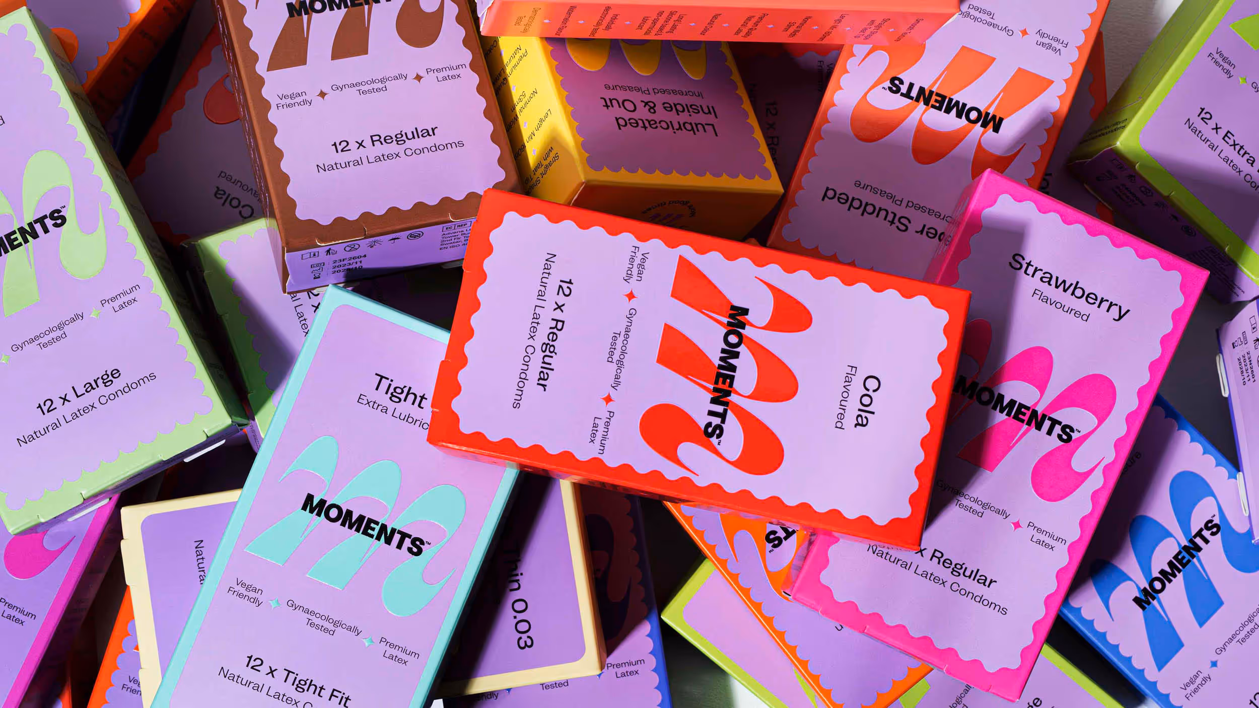 Assorted colorful boxes of Moments natural latex condoms in various sizes and flavors stacked together.