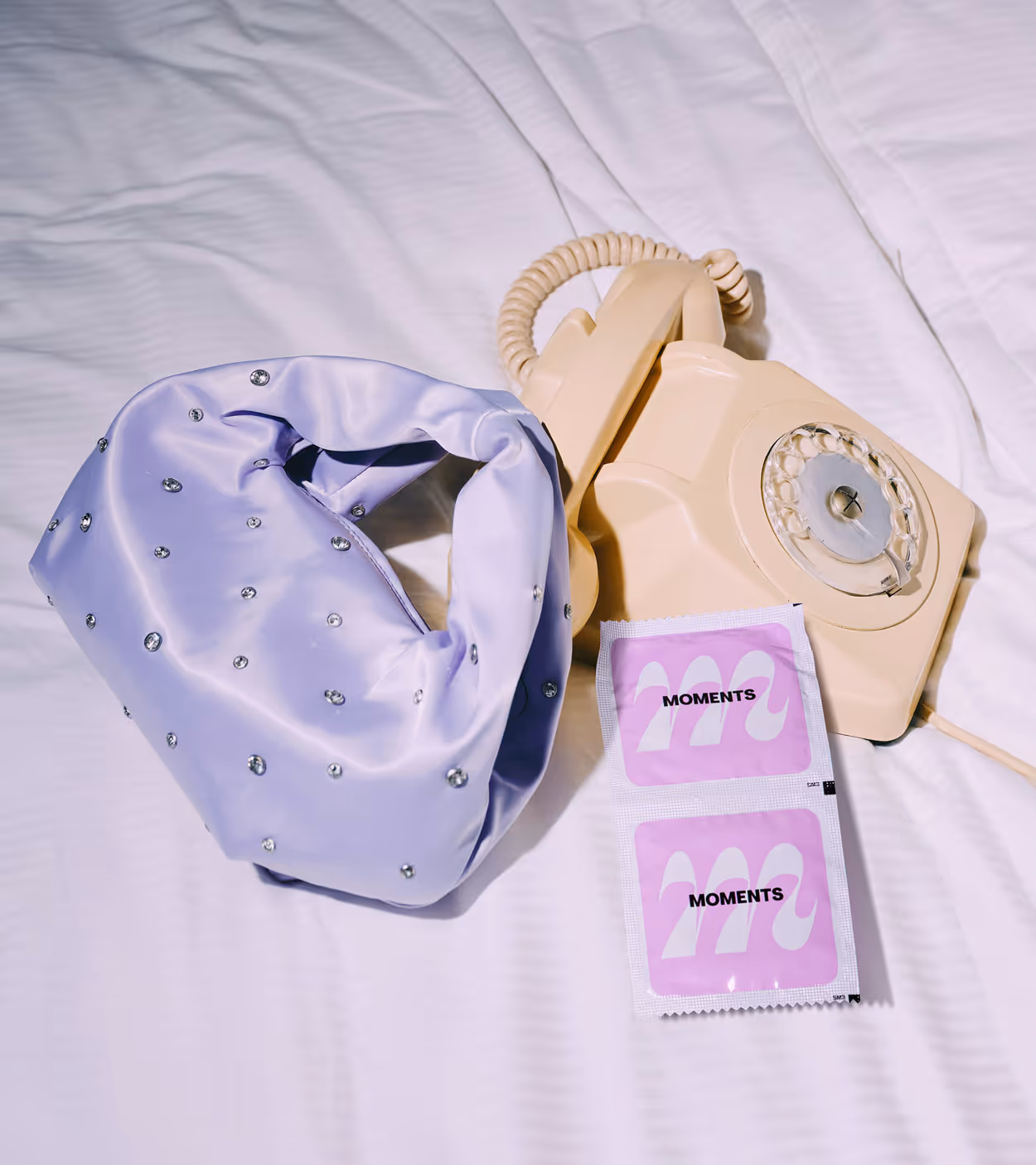 Beige rotary telephone, lavender satin handbag with rhinestones, and two pink 'Moments' condom packets on a white bedsheet.