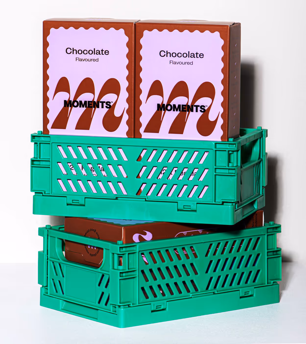 Two chocolate-flavored Moments snack boxes stacked in green plastic crates against a white background.