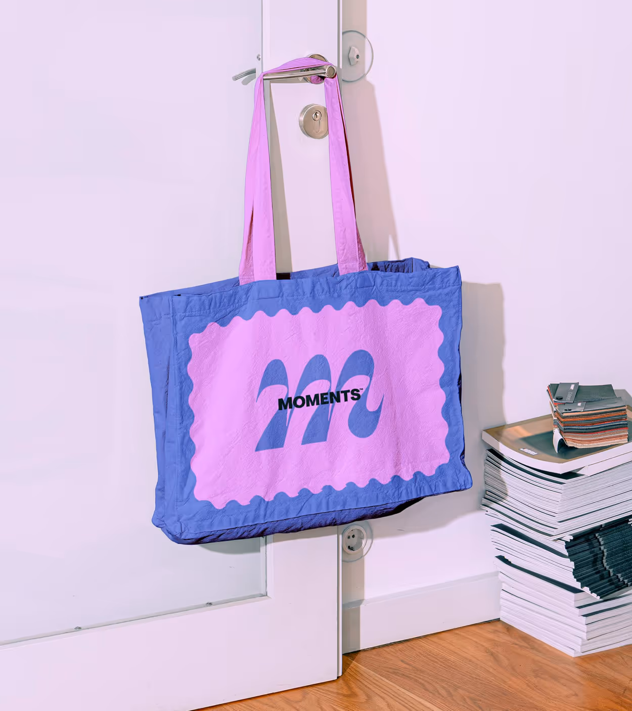 Blue and pink tote bag with wavy pink design and word 'MOMENTS' hanging on a silver door handle.