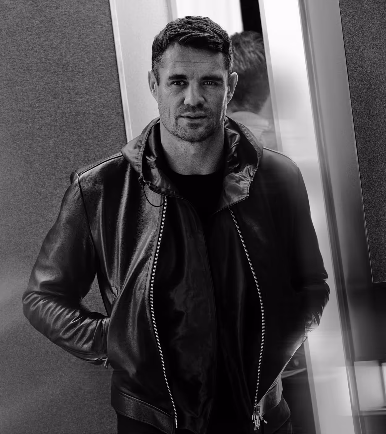 Black and white portrait of a man wearing a leather jacket standing with hands in pockets near a reflective surface.