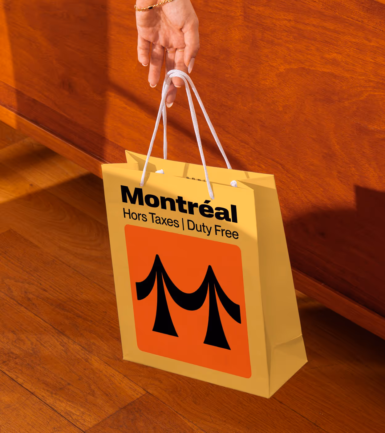 Hand holding a yellow shopping bag with white handles that reads 'Montréal Hors Taxes | Duty Free' and features a black stylized bridge on an orange square.