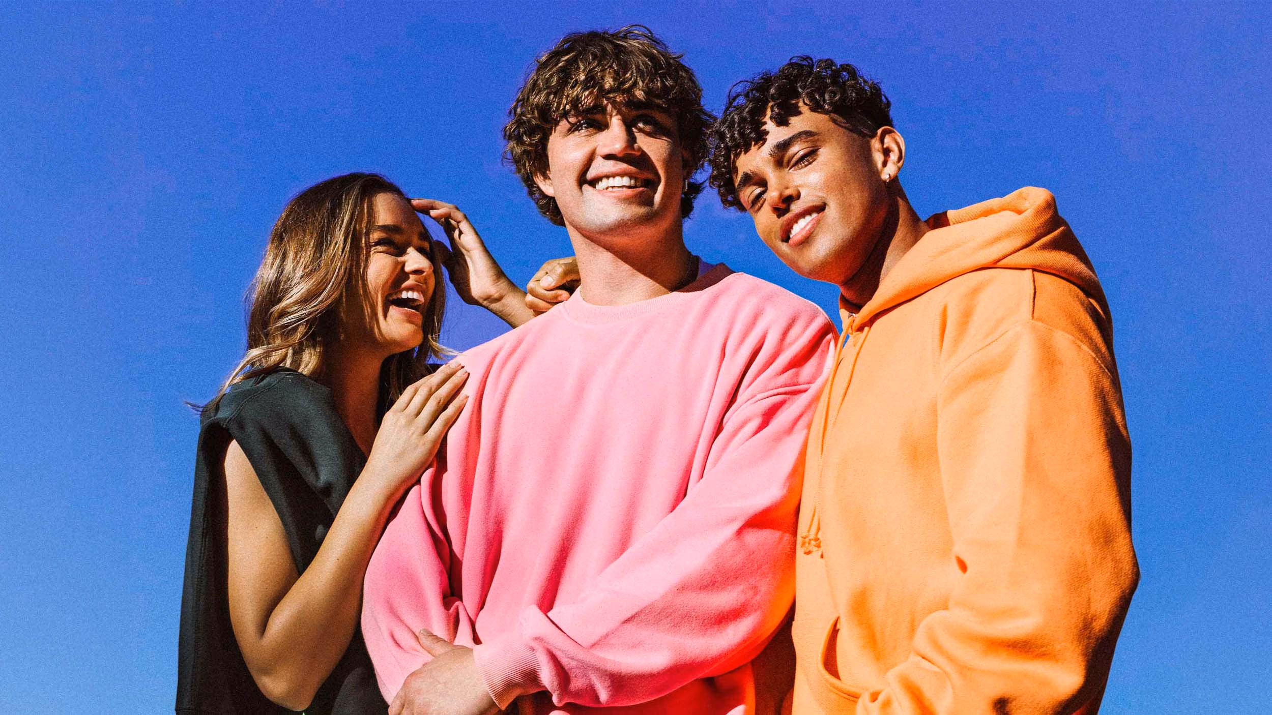 Three young adults smiling and standing close together under clear blue sky, one in a pink sweatshirt, one in an orange hoodie, and one in a black sleeveless top.