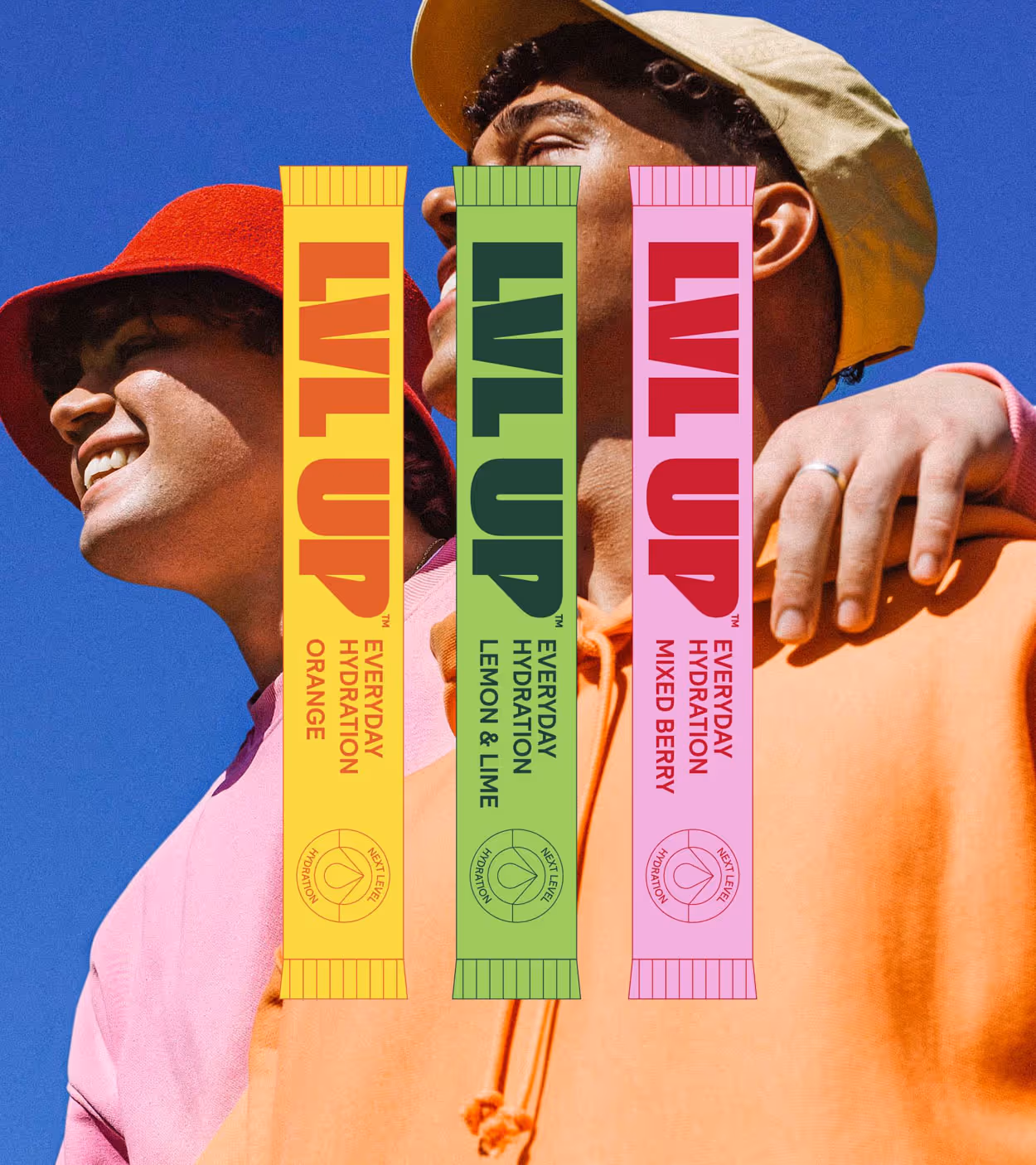 Two young men smiling outdoors wearing colorful hats and hoodies, with three LVL UP hydration packets (orange, lemon & lime, mixed berry) overlaid in front.